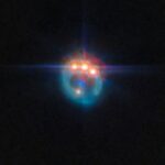 Una piccola immagine di una galassia distorta dalla lente gravitazionale in un anello fioco. Nella parte superiore dell'anello ci sono tre punti molto luminosi con punte di diffrazione che fuoriescono da essi, uno accanto all'altro: in realtà si tratta di copie di un singolo quasar nella galassia, duplicate dalla lente gravitazionale. Al centro dell'anello, la galassia ellittica che esegue la lente appare come un piccolo punto blu.