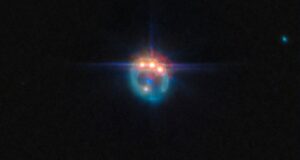 Una piccola immagine di una galassia distorta dalla lente gravitazionale in un anello fioco. Nella parte superiore dell'anello ci sono tre punti molto luminosi con punte di diffrazione che fuoriescono da essi, uno accanto all'altro: in realtà si tratta di copie di un singolo quasar nella galassia, duplicate dalla lente gravitazionale. Al centro dell'anello, la galassia ellittica che esegue la lente appare come un piccolo punto blu.