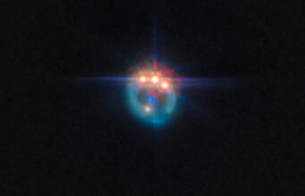 L’Anello Prezioso Creato da un Quasar Una piccola immagine di una galassia distorta dalla lente gravitazionale in un anello fioco. Nella parte superiore dell'anello ci sono tre punti molto luminosi con punte di diffrazione che fuoriescono da essi, uno accanto all'altro: in realtà si tratta di copie di un singolo quasar nella galassia, duplicate dalla lente gravitazionale. Al centro dell'anello, la galassia ellittica che esegue la lente appare come un piccolo punto blu.