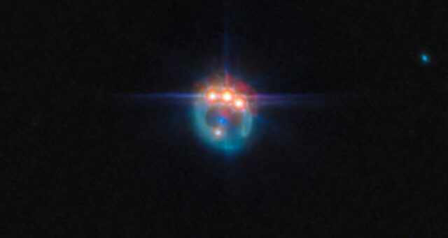 Una piccola immagine di una galassia distorta dalla lente gravitazionale in un anello fioco. Nella parte superiore dell'anello ci sono tre punti molto luminosi con punte di diffrazione che fuoriescono da essi, uno accanto all'altro: in realtà si tratta di copie di un singolo quasar nella galassia, duplicate dalla lente gravitazionale. Al centro dell'anello, la galassia ellittica che esegue la lente appare come un piccolo punto blu.